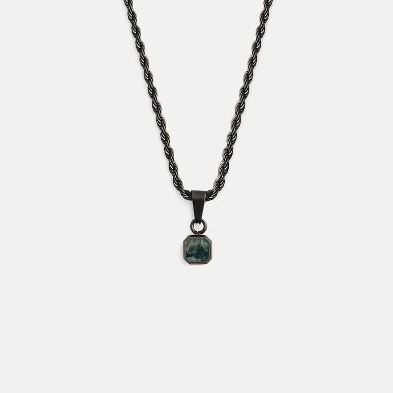 May include: A black necklace with a square pendant. The pendant features a dark green stone set in a black metal frame. The chain has a rope-like design. The necklace is displayed against a white background.