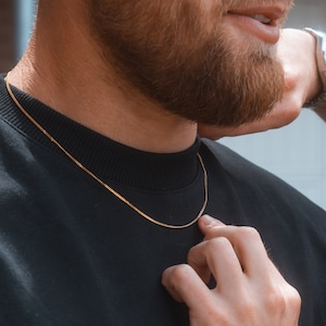 May include: A gold chain necklace worn by a person with a beard. The chain is thin and has a box link style.
