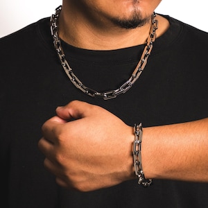 May include: A silver-colored chain necklace and bracelet set. The necklace is around the neck, and the bracelet is on the wrist. Both pieces have a rectangular link design. The person is wearing a black shirt.