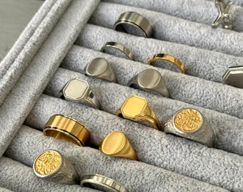 May include: A collection of various rings displayed in a gray velvet jewelry tray. The rings are made of gold and silver metals, featuring different designs such as signet, textured, and band styles. The rings are in various shapes, including oval, square, and round.