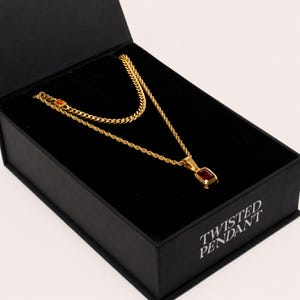 May include: A gold-toned layered necklace set featuring a rope chain with a rectangular red gemstone pendant and a secondary curb chain. The jewelry is displayed inside a black presentation box with the text TWISTED PENDANT printed on the side.