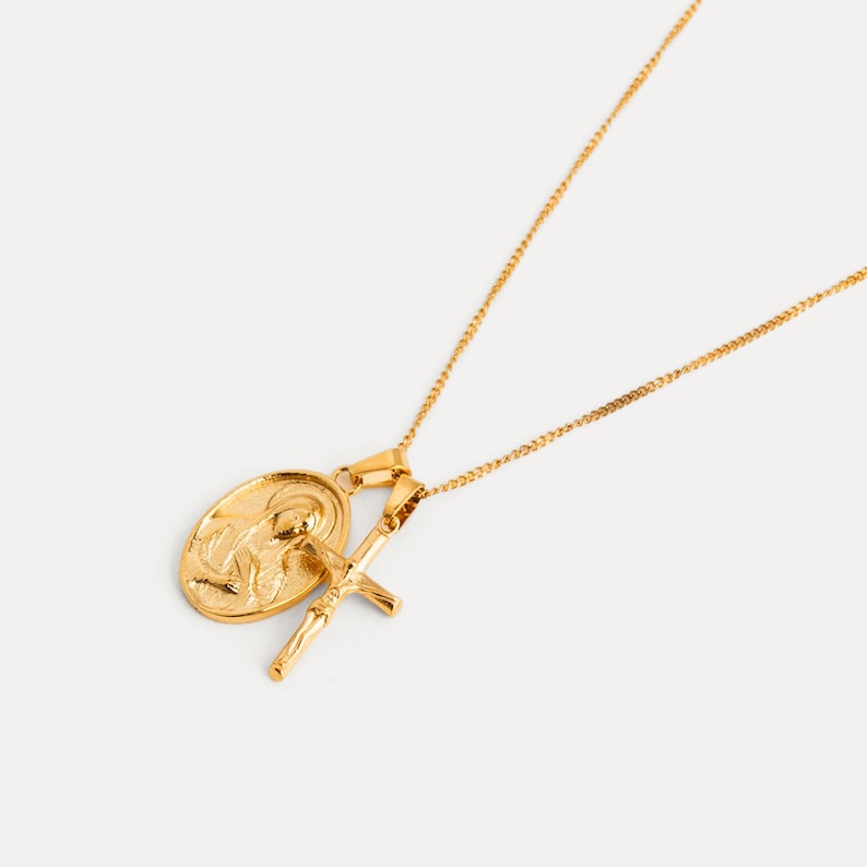 May include: Gold-colored necklace featuring two pendants: an oval medallion with a religious figure and a crucifix. The pendants hang from delicate chains, creating a layered effect. The jewelry is set against a plain background.