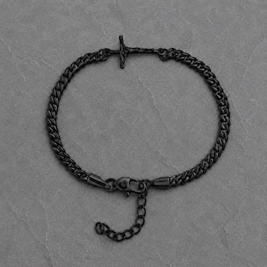 May include: A matte black metal chain bracelet featuring a small crucifix charm integrated into the links. The bracelet includes a lobster claw clasp and an adjustable extender chain with circular links, providing a secure and customisable fit.