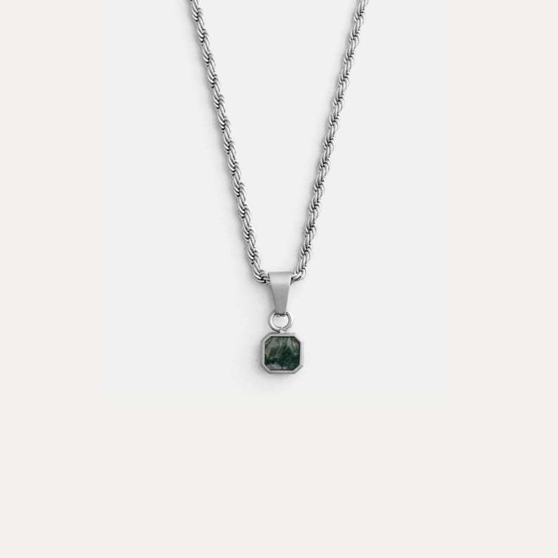 May include: A silver-toned necklace with a square pendant. The pendant features a dark green stone set in a silver frame. The chain is a twisted rope design. The necklace is displayed against a white background.
