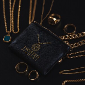 May include: A black leather pouch with a gold zipper and the text "Twisted Pendant" printed on the front. The pouch is surrounded by gold chains and rings.