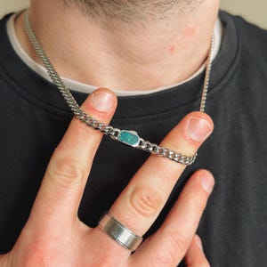 May include: A silver chain necklace with a turquoise-colored rectangular pendant. The necklace is a curb chain style. A silver ring is on the finger. The person is wearing a black shirt.