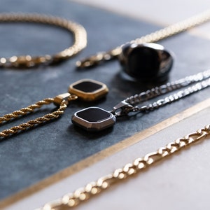 May include: An assortment of men's jewelry, including gold and silver necklaces, a bracelet, and a black stone ring. The necklaces feature geometric pendants with black insets. The jewelry is displayed on a marble surface.