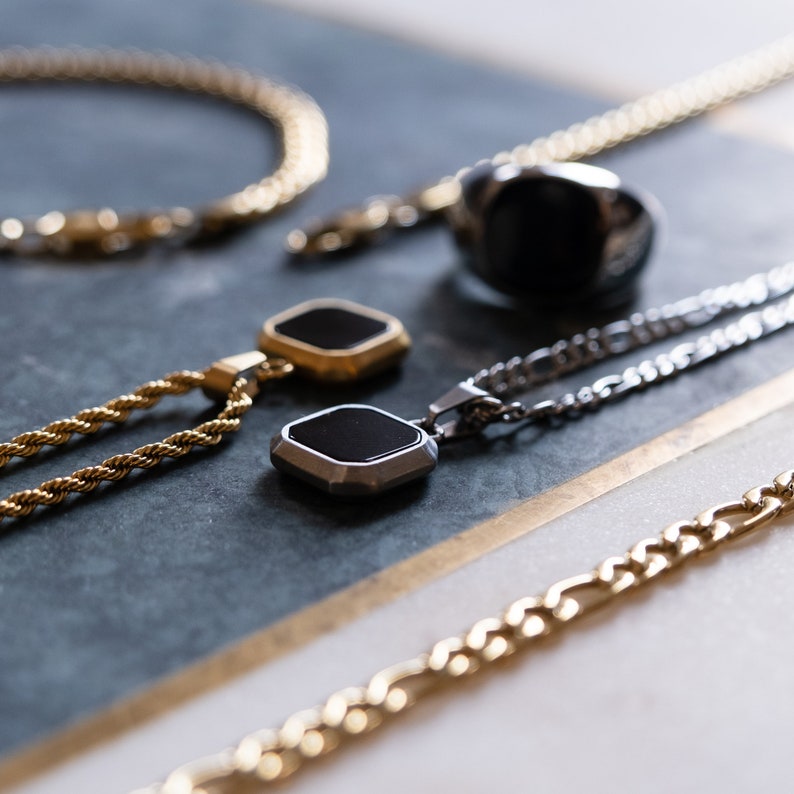 May include: An assortment of men's jewelry, including gold and silver necklaces, a bracelet, and a black stone ring. The necklaces feature geometric pendants with black insets. The jewelry is displayed on a marble surface.