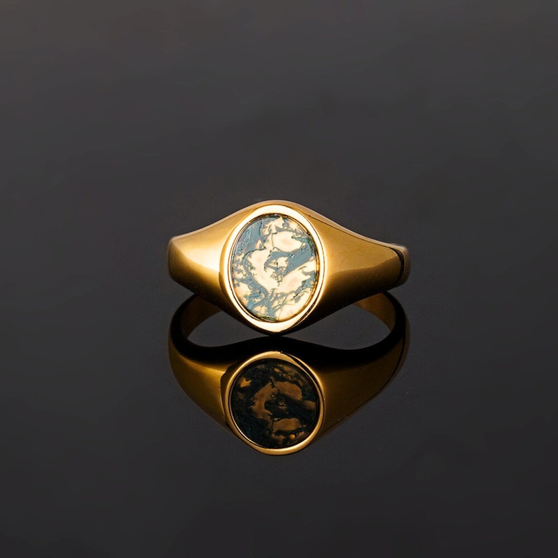 May include: A gold-toned signet ring featuring an oval-shaped inlay with a marbled, moss-like green and cream pattern. The ring has a polished, smooth metal band and a reflective surface that mirrors the design of the stone setting below it.