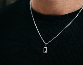 May include: Silver pendant necklace with a rectangular black stone. The pendant is surrounded by a textured silver frame and hangs from a silver rope chain. The necklace is displayed against a black background.