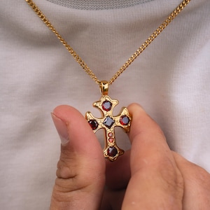 Può includere: Collana con pendente a croce color oro con catena a maglia. La croce presenta pietre preziose rosse e rosso scuro. La collana è tenuta da una persona che indossa una camicia bianca. È un simbolo religioso.