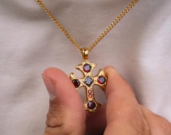 Op de afbeelding: Goudkleurige kruisvormige hangerketting met een gourmetketting. Het kruis heeft rode en donkerrode edelstenen. De ketting wordt vastgehouden door een persoon die een wit shirt draagt. Het kruis is een religieus symbool.
