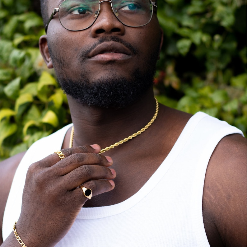 Puede incluir: Primer plano de una persona que lleva un top blanco sin mangas, un collar de cadena de cuerda dorada y un anillo de oro con una piedra negra. La persona lleva gafas y tiene barba.