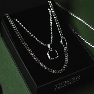 May include: A silver-toned necklace set with a black square pendant, displayed in a black box. The set includes a rope chain and a curb chain. The box has the words "TWISTED PENDANT" printed on it.