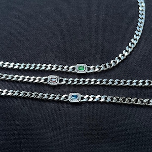 May include: Three silver chain necklaces with rectangular gemstone accents. One necklace features a green gemstone, another a red gemstone, and the third a blue gemstone. The necklaces are displayed on a dark background.
