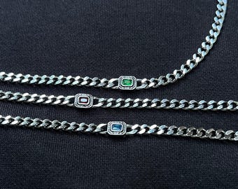 May include: Three silver chain necklaces with rectangular gemstone accents. One necklace features a green gemstone, another a red gemstone, and the third a blue gemstone. The necklaces are displayed on a dark background.