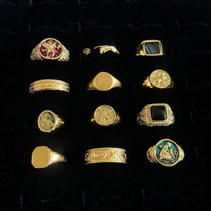 Könnte beinhalten: Eine Sammlung von zwölf goldfarbenen Ringen, die in einem Raster angeordnet sind. Die Ringe weisen verschiedene Designs auf, darunter schlichte Bänder, Siegelringe, gravierte Muster sowie runde oder quadratische Fassungen mit roten, schwarzen, grünen und goldfarbenen Einlagen.