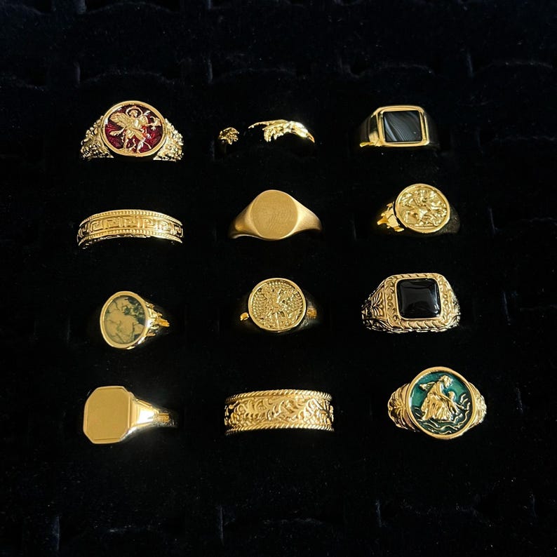 May include: A collection of twelve gold-toned rings arranged in a grid. The rings feature various designs including solid bands, signet styles, engraved patterns, and circular or square settings with red, black, green, and gold-colored inlays.