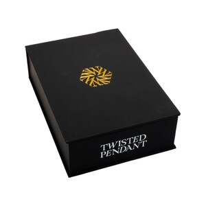 May include: A black rectangular box with a gold geometric design on the lid. The words "TWISTED PENDANT" are printed in white on the front of the box. The box is closed and appears to be made of cardboard.