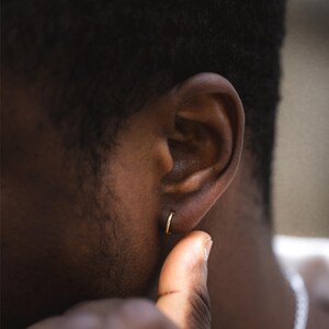 Puede incluir: Un peque&ntilde;o pendiente de aro dorado se lleva en la oreja de una persona de piel oscura. El pendiente tiene un dise&ntilde;o simple y circular. La mano de la persona es visible, tocando la oreja.