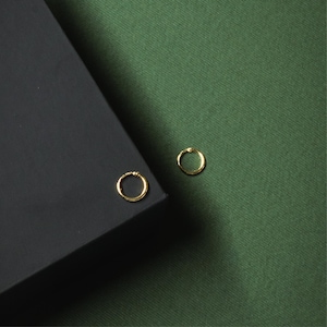 Puede incluir: Dos pequeños pendientes de aro dorados. Un pendiente descansa sobre una superficie negra, mientras que el otro se encuentra sobre un fondo de tela verde. Los pendientes tienen un diseño simple y minimalista.