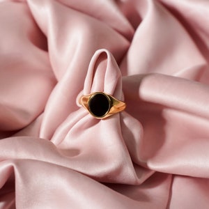 Op de afbeelding: Een gouden zegelring met een zwarte ovale steen. De ring is geplaatst tegen een achtergrond van zachte, roze satijnen stof, wat een luxe en elegante uitstraling cre&euml;ert. Het ontwerp van de ring is eenvoudig maar verfijnd.