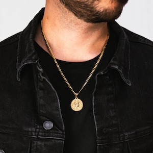 Puede incluir: Un collar de cadena dorada con un colgante redondo que presenta un diseño detallado. El collar lo lleva una persona que lleva una camisa negra y una chaqueta vaquera negra.