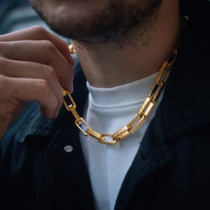 Puede incluir: Un collar de cadena dorado con eslabones rectangulares. El collar es sostenido por una mano, mostrando su diseño y textura detallados. La cadena se lleva sobre un cuello alto blanco y una chaqueta negra.
