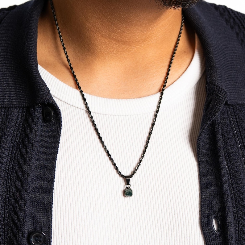 May include: A black rope chain necklace with a small, square, dark green pendant. The necklace is worn over a white ribbed undershirt and a dark blue cardigan.