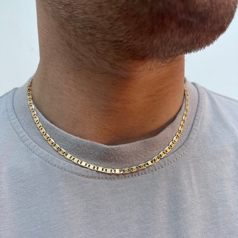 Może przedstawiać: Złoty naszyjnik z łańcuszkiem w stylu marynarskim. Łańcuszek nosi osoba w jasnoszarym t-shircie.