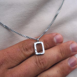 Puede incluir: Collar plateado con un colgante rectangular. El colgante tiene un aspecto texturizado y martillado, colgado de una cadena fina. La imagen muestra el collar sobre un fondo neutro.