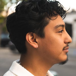 Puede incluir: Perfil lateral de una persona con cabello oscuro y rizado y un peque&ntilde;o pendiente de aro plateado. La persona tiene bigote y barba corta. Lleva una camisa blanca texturizada con un collar de cadena plateada.