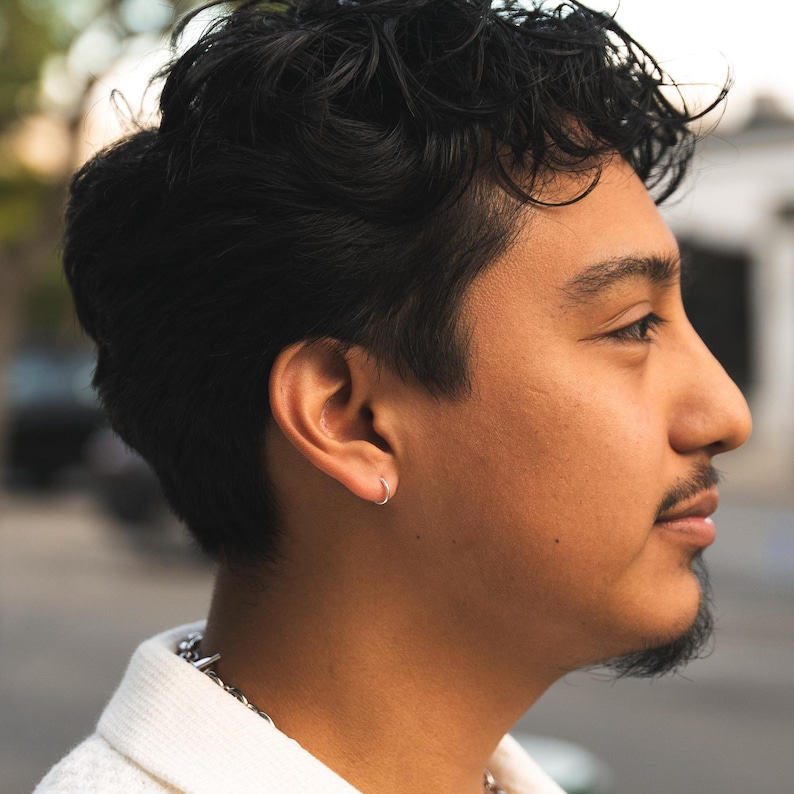 Puede incluir: Perfil lateral de una persona con cabello oscuro y rizado y un peque&ntilde;o pendiente de aro plateado. La persona tiene bigote y barba corta. Lleva una camisa blanca texturizada con un collar de cadena plateada.