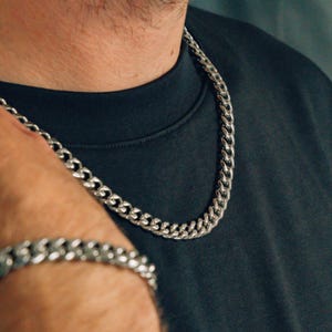 Może przedstawiać: Srebrny naszyjnik i bransoletka z łańcucha. Naszyjnik jest gruby i ma wzór łańcucha typu curb. Bransoletka jest cieńsza i ma podobny wzór.