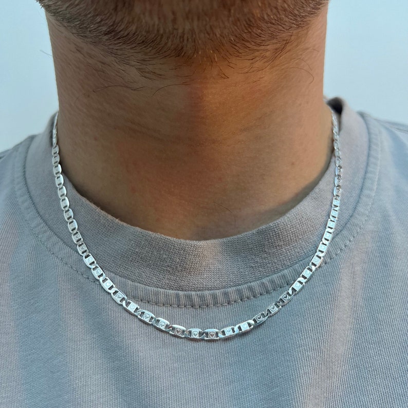 May include: A silver chain necklace with a mariner link design. The chain is worn by a person with a grey shirt.