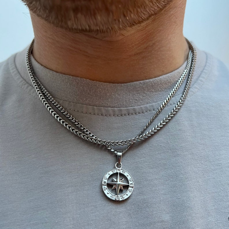 May include: A silver-colored necklace with a compass pendant. The pendant features a star design within a circular frame, suspended from a wheat chain. The necklace is layered over a second, similar chain. The person is wearing a gray t-shirt.