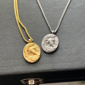 May include: Two necklaces with oval pendants featuring a detailed profile of a man wearing a helmet. One necklace has a gold chain and pendant, the other has a silver chain and pendant.