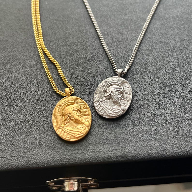 May include: Two necklaces with oval pendants featuring a detailed profile of a man wearing a helmet. One necklace has a gold chain and pendant, the other has a silver chain and pendant.