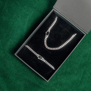 Pode incluir: Um conjunto de colar e pulseira de corrente prateada com detalhes pretos, apresentado numa caixa preta com exterior cinzento. As joias são exibidas numa superfície de veludo preto, sobre um fundo verde.