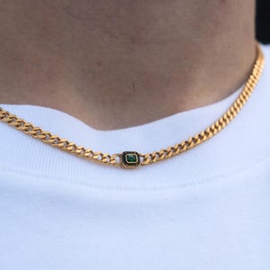 May include: Gold-colored chain necklace with a rectangular green gemstone pendant. The necklace is a curb chain style, and the pendant is set in a black frame. The necklace is worn against a white shirt.