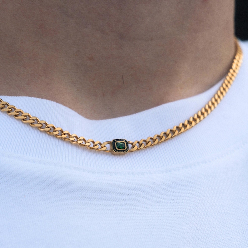 May include: Gold-colored chain necklace with a rectangular green gemstone pendant. The necklace is a curb chain style, and the pendant is set in a black frame. The necklace is worn against a white shirt.