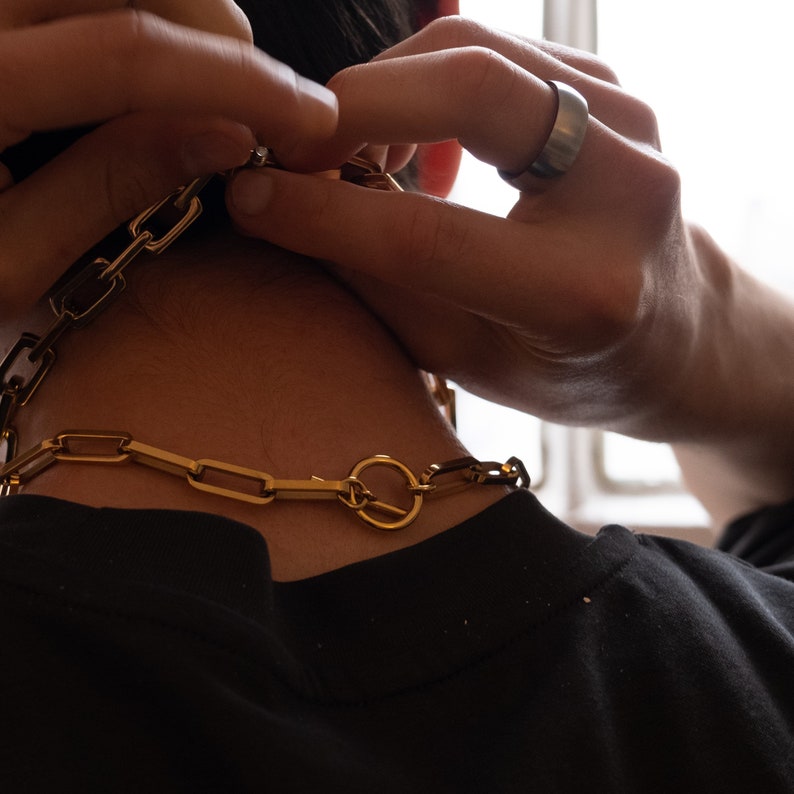 May include: A gold-colored chain necklace with large rectangular links and a circular clasp. The necklace is being fastened around the neck. A silver ring is visible on the hand.