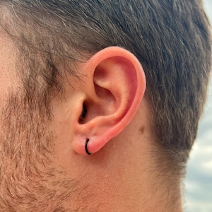 May include: A close-up of a person's ear with a small, black hoop earring. The ear is pinkish-red, and the skin around it is light-toned. The person's hair is dark and slightly out of focus in the background.