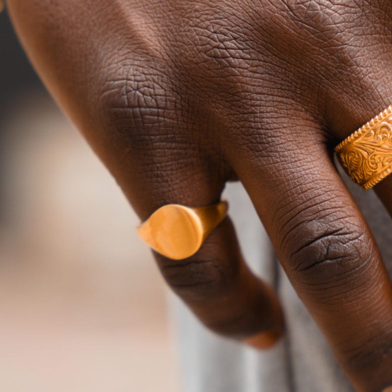 May include: A gold signet ring with a smooth, polished, circular face on the ring finger and a wider gold band featuring an intricate, embossed floral or scrollwork pattern on the adjacent finger, both displaying a warm, metallic yellow finish.