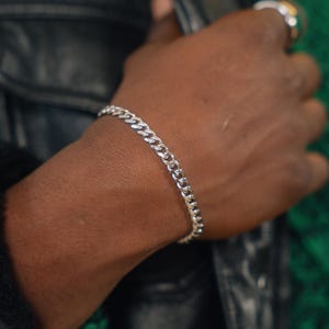 Op de afbeelding: Een zilveren gourmet armband wordt om een pols gedragen. De armband is gemaakt van in elkaar grijpende schakels en heeft een glanzende, metalen afwerking. De achtergrond omvat een zwarte leren jas en een groen patroon.