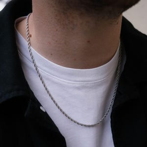 May include: A silver rope chain necklace worn by a person wearing a white t-shirt and a black jacket.