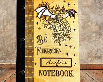 May include: A wooden notebook with a dragon design. The cover is light brown with a laser-cut dragon, floral, and star details. The text "Be Fierce" and "Aoife's Notebook" are also laser-cut. The spine is black with gold rivets.