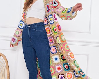 May include: A long, crocheted cardigan with a colorful floral pattern. The cardigan is made of a beige yarn and features a variety of bright colors, including pink, blue, green, yellow, and orange. The cardigan is worn over a white crop top and blue jeans.