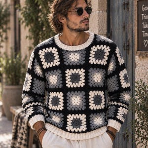 Op de afbeelding: Een trui met lange mouwen voorzien van een gehaakt granny square patroon in tinten zwart, antracietgrijs, lichtgrijs en gebroken wit. Het kledingstuk heeft een ronde halslijn en geribbelde boorden aan de mouwen en zoom in effen gebroken wit.