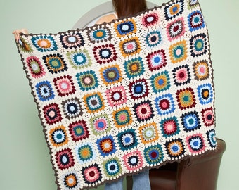 May include: A colourful crocheted throw blanket with a granny square pattern in shades of red, blue, green, yellow, and brown. The blanket is made with a white background and has a brown border.